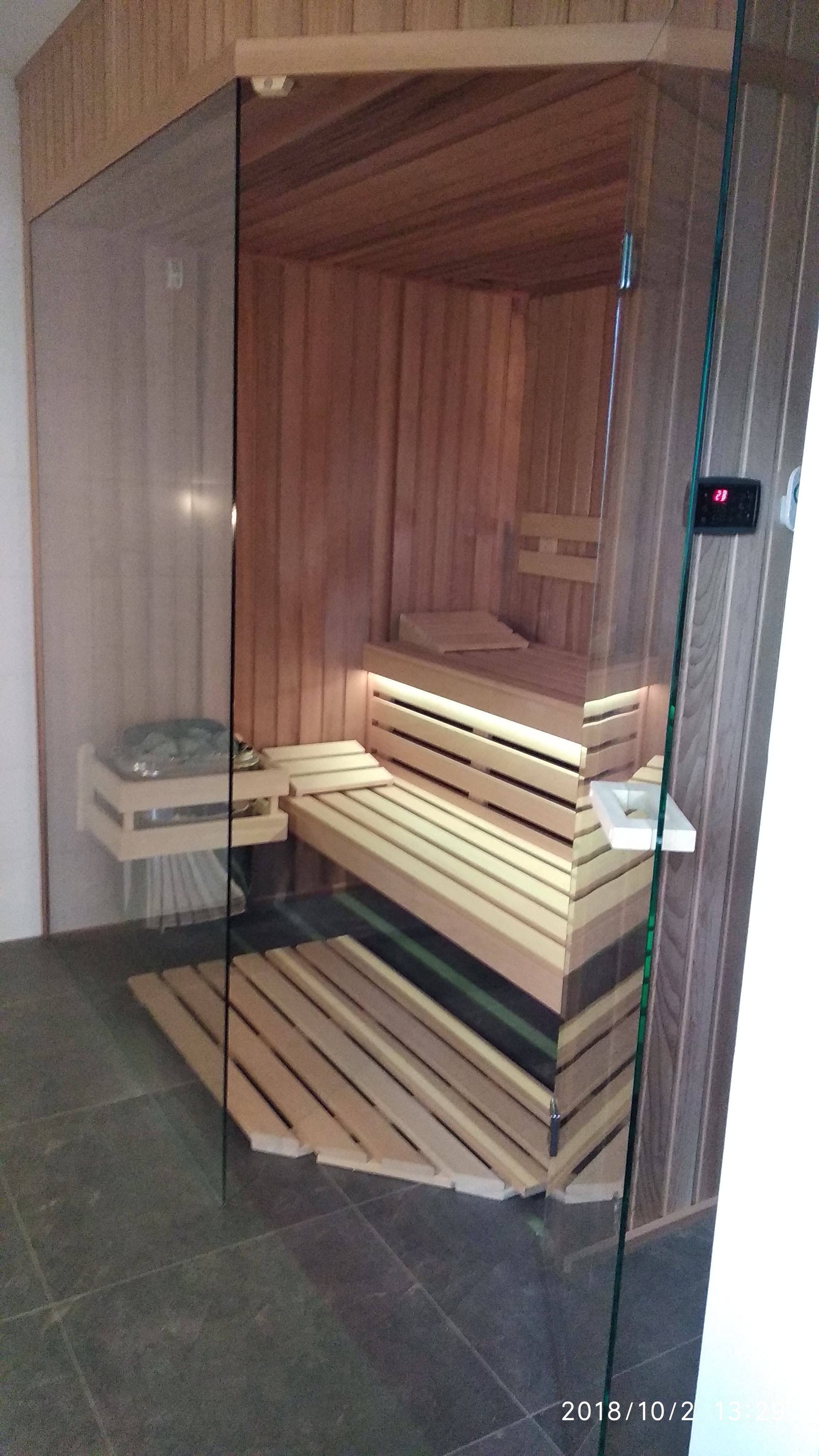 Wnętrze sauny z jasnego drewna z przeszklonymi ścianami, widoczne ławy, podświetlenie LED i kamienie na półce.