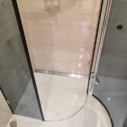 Narożna kabina prysznicowa z czarnymi profilami, beżowe płytki z motywem roślinnym, odpływ liniowy w podłodze, widoczne fragmenty podłogi i ściany w trakcie montażu.