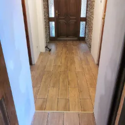 Korytarz w trakcie układania podłogi z płytek imitujących drewno, widoczne krzyżyki dystansowe, fragment ściany z dekoracyjnym kamieniem i drewniane drzwi wejściowe.