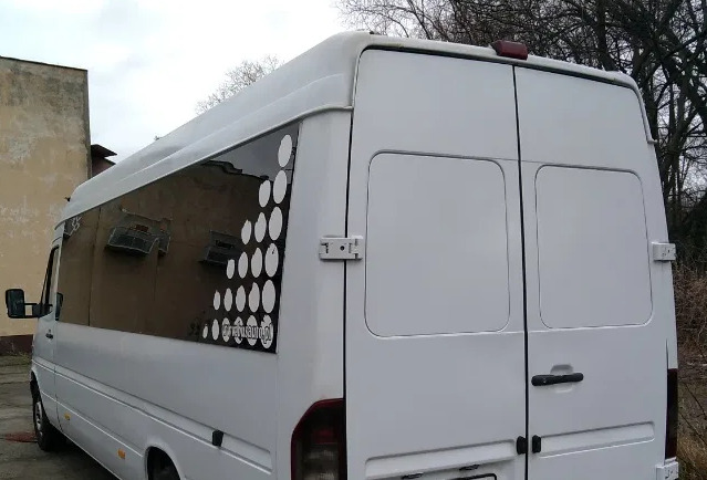 Biały bus z ciemnymi szybami i naklejką z białych kropek, zaparkowany przy budynku w pochmurny dzień.