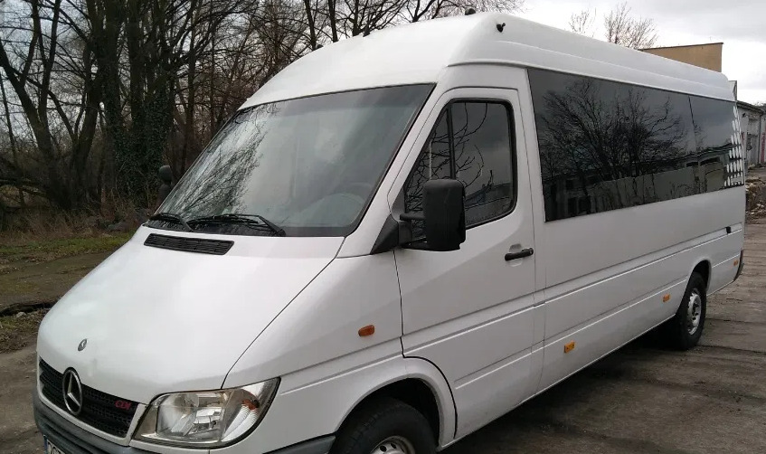 Biały bus Mercedes-Benz Sprinter z przyciemnianymi szybami, zaparkowany na betonowym placu z drzewami w tle.
