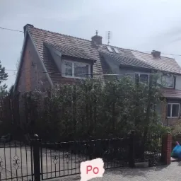 Dwupiętrowy dom z czerwonej cegły z brązowym dachem, widoczny zza wysokiego żywopłotu i czarnego metalowego ogrodzenia, z napisem 'Po' na pierwszym planie.