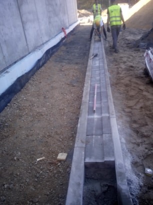 Montaż korytka odwadniającego z kostki betonowej na placu budowy, widoczny poziomica i pracownicy w oddali.