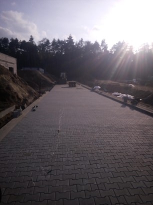 Ułożona szara kostka brukowa na długim podjeździe w Nowym Kozłowie, z widocznym sprzętem budowlanym w oddali i skarpą po lewej stronie.