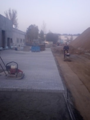 Prace brukarskie w toku: ubijanie podłoża pod kostkę brukową za pomocą zagęszczarki, widoczne ułożone fragmenty kostki i przygotowane zapasy na paletach w tle, pochmurny dzień.