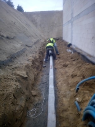 Dwóch pracowników w żółtych kamizelkach montuje długą, metalową rynnę odwadniającą w wykopie ziemnym, widoczne kable i niebieskie rury.