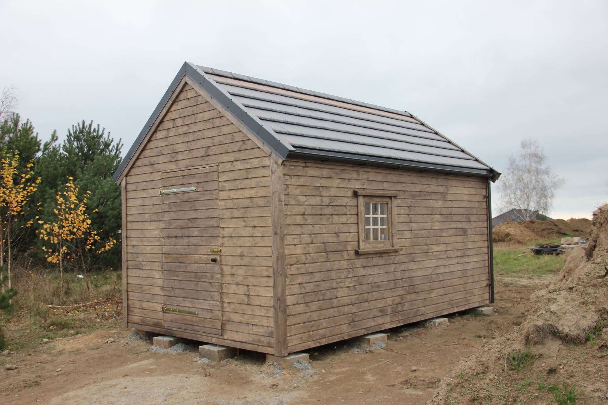 Mały, drewniany domek z ciemną dachówką, osadzony na betonowych bloczkach. Widoczne drzwi, okno i fragment otoczenia z zielenią i ziemią.