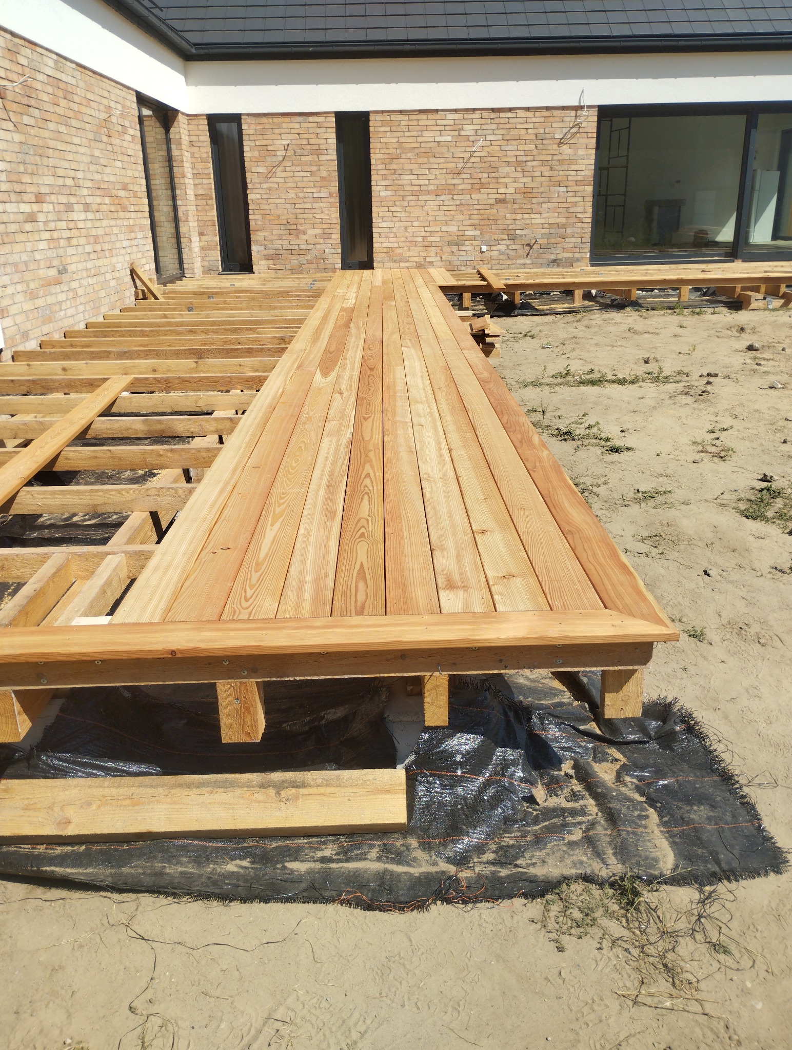 Częściowo ułożony drewniany taras przy domu z cegły. Widoczne deski, konstrukcja nośna i czarna geowłóknina. Etap budowy tarasu.