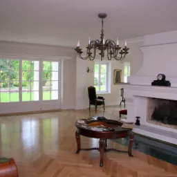 Przestronny salon z parkietem w jodełkę, dużym kominkiem z ozdobną obudową, żyrandolem i widokiem na ogród przez przeszklone drzwi.