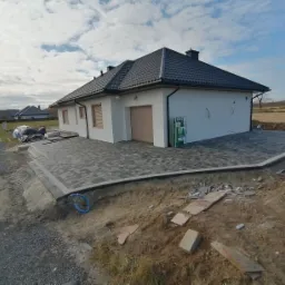 Ułożona kostka brukowa w szarych odcieniach przed nowoczesnym, białym domem z garażem, widok na posesję w trakcie prac wykończeniowych.