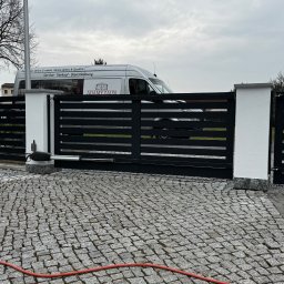 ADAMY.ZAUN - Automatyczna brama wjazdowa z poziomych, czarnych profili, osadzona na białych słupkach z szarymi nakryciami, widok z kostki brukowej w Bieżycach, z samochodem dostawczym firmy w tle.