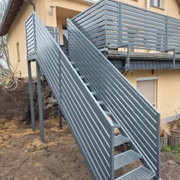 ADAMY.ZAUN - Zewnętrzne schody metalowe z balustradą z poziomych profili, prowadzące na taras domu w trakcie montażu, widoczny wykop w ziemi i betonowe donice.
