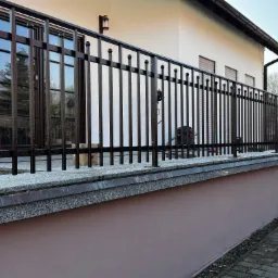 Metalowa balustrada balkonowa w kolorze ciemnego brązu z kulkami na szczycie pionowych prętów, zamontowana na granitowym murku, z widokiem na okna budynku w tle.