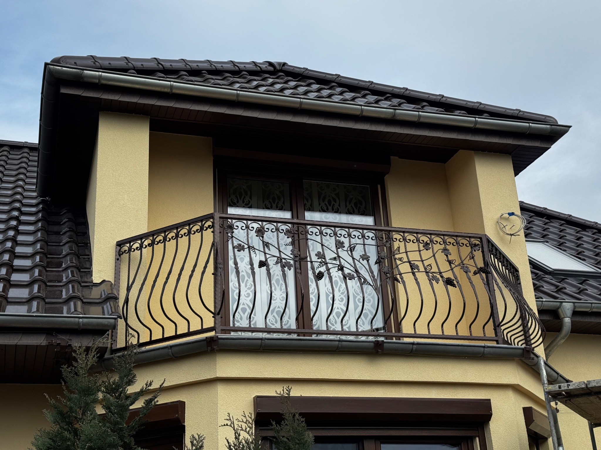 Balkon z ozdobną, metalową balustradą z motywem winorośli na tle beżowej elewacji domu z brązową dachówką.