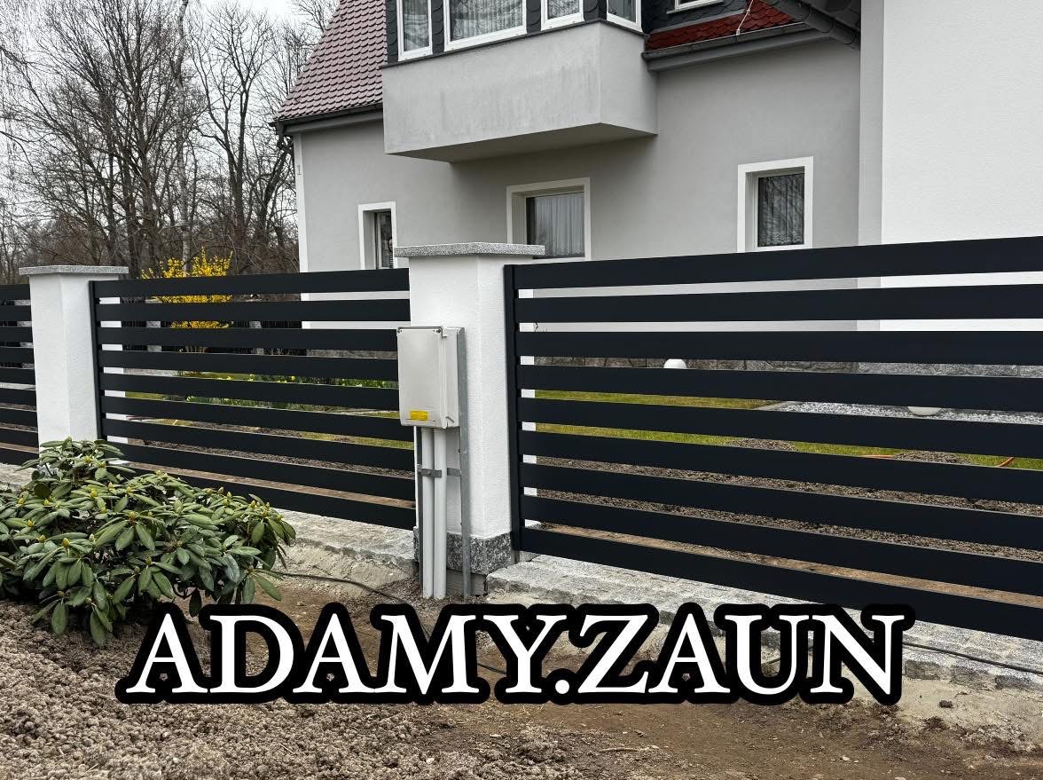 Nowoczesne, poziome, czarne przęsła ogrodzeniowe z białymi słupkami na tle szarego domu z brązowym dachem, widoczna skrzynka elektryczna na słupku.