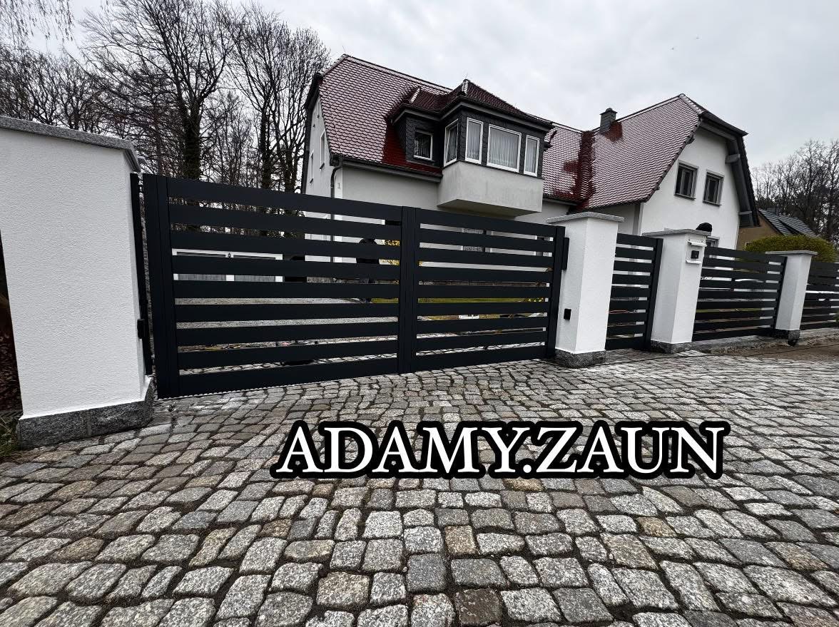 Nowoczesna, pozioma brama wjazdowa i przęsła ogrodzeniowe z czarnych, metalowych profili, osadzone na białych, murowanych słupkach; podjazd wyłożony kostką brukową, w tle dom jednorodzinny...