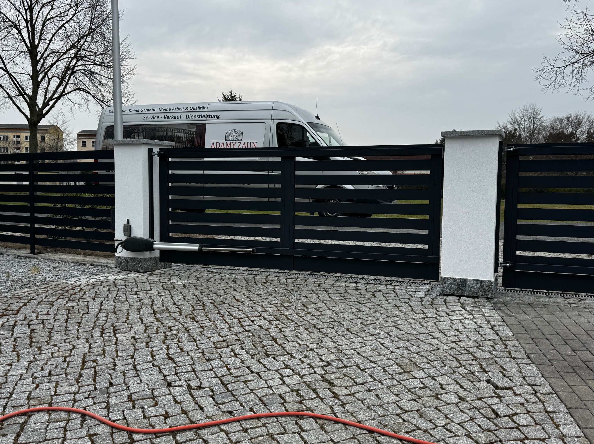 Automatyczna brama wjazdowa z poziomych, czarnych profili, osadzona na białych słupkach z szarymi nakryciami, widok z kostki brukowej w Bieżycach, z samochodem dostawczym firmy w tle.