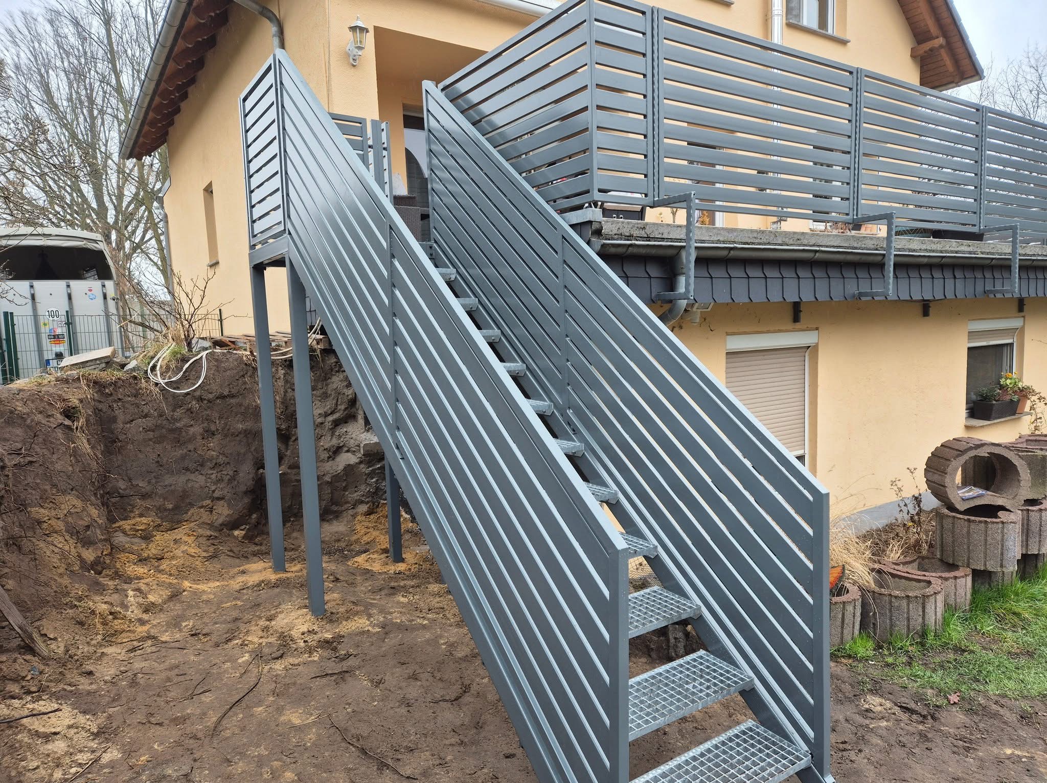 Zewnętrzne schody metalowe z balustradą z poziomych profili, prowadzące na taras domu w trakcie montażu, widoczny wykop w ziemi i betonowe donice.