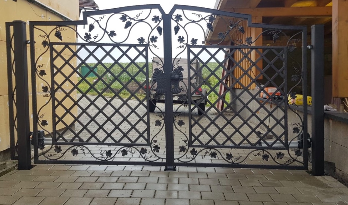 Dwuskrzydłowa, czarna brama wjazdowa z motywem winogron i liści, kratą w romby oraz ozdobnym orłem pośrodku, zamontowana na posesji z brukowanym podjazdem.