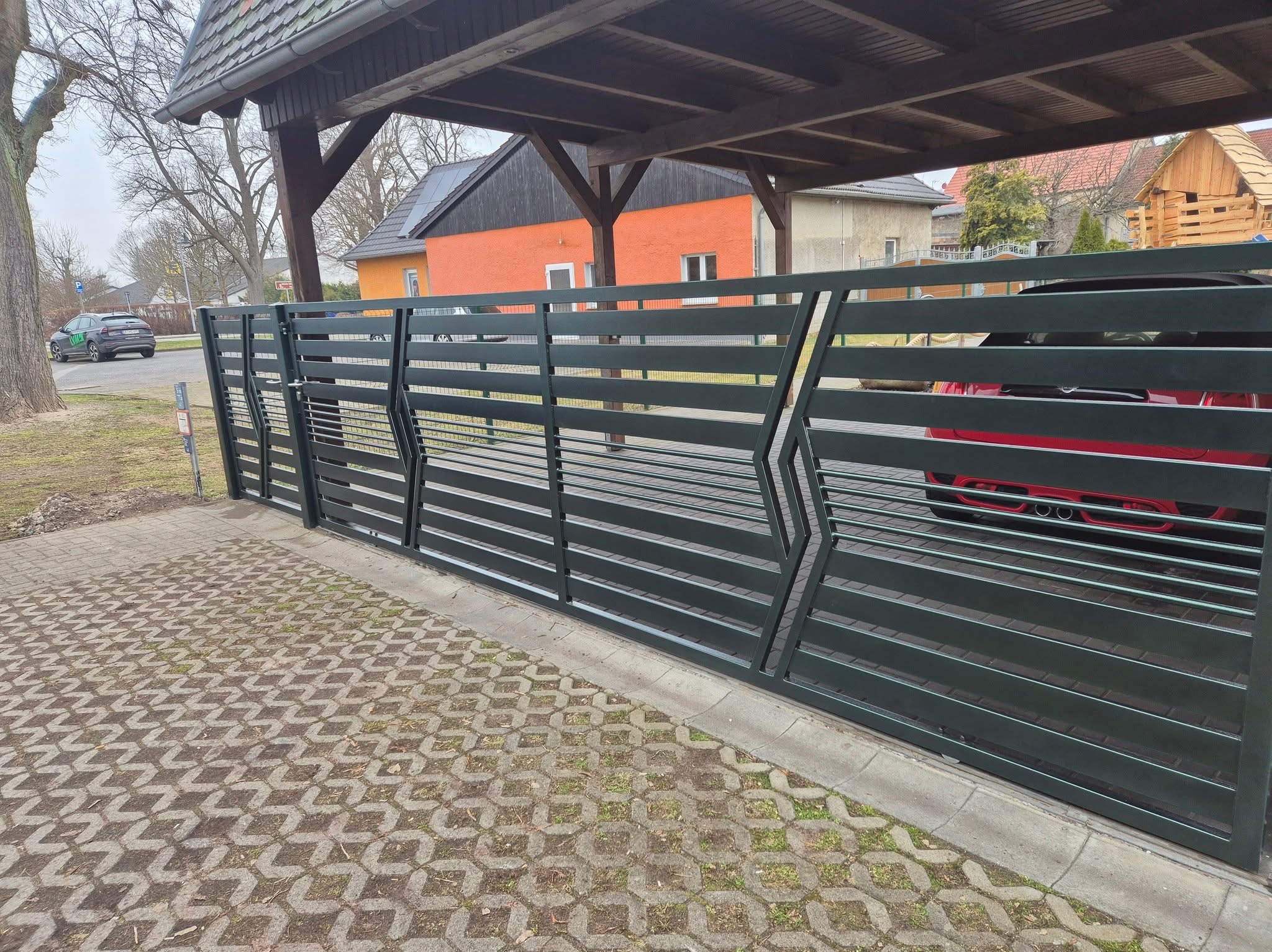 Nowoczesna, pozioma brama metalowa w kolorze grafitowym z geometrycznym wzorem w kształcie strzałek, prowadząca na podjazd wyłożony ażurową kostką; w tle widoczny czerwony samochód i domy.
