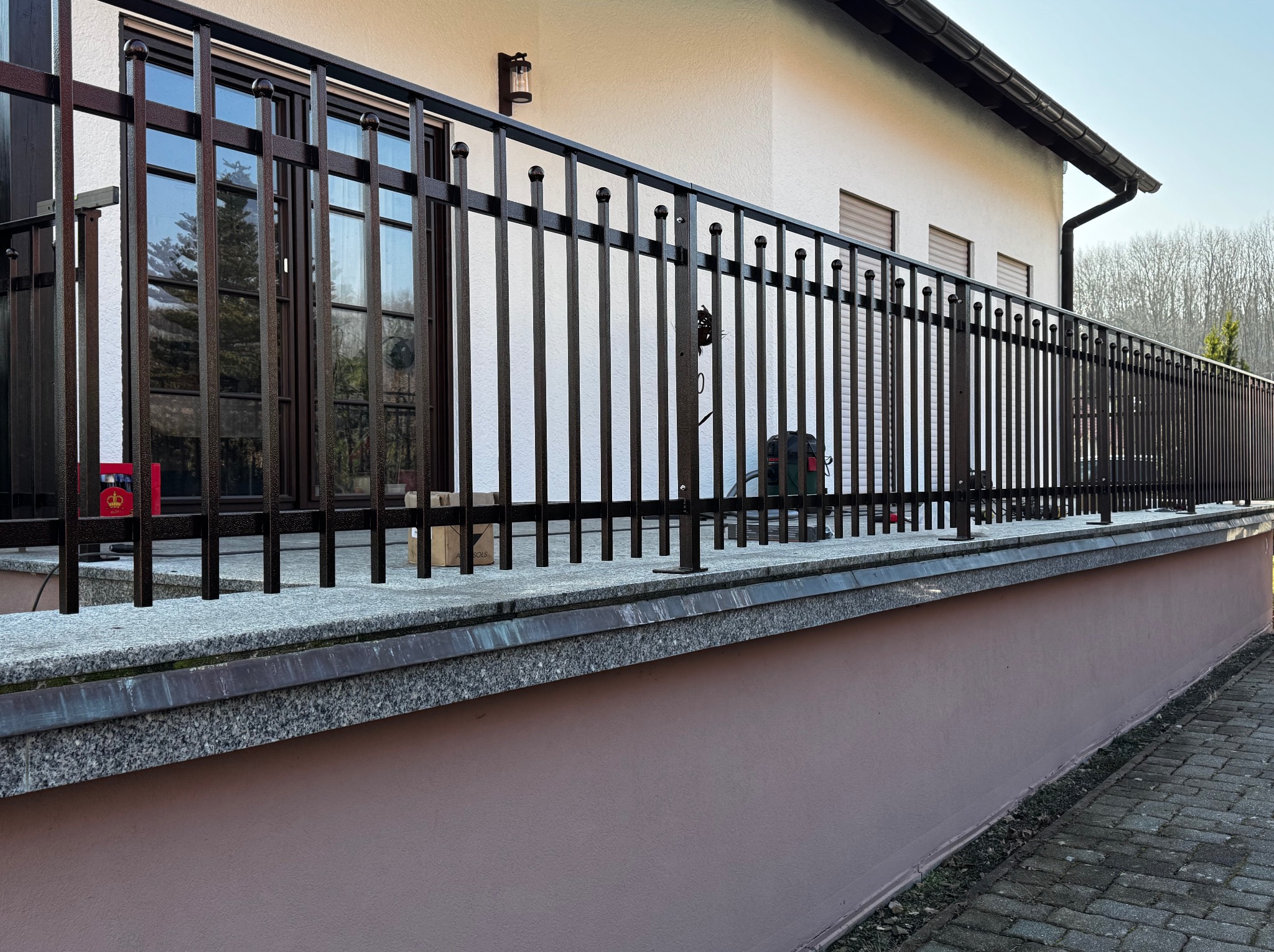 Metalowa balustrada balkonowa w kolorze ciemnego brązu z kulkami na szczycie pionowych prętów, zamontowana na granitowym murku, z widokiem na okna budynku w tle.