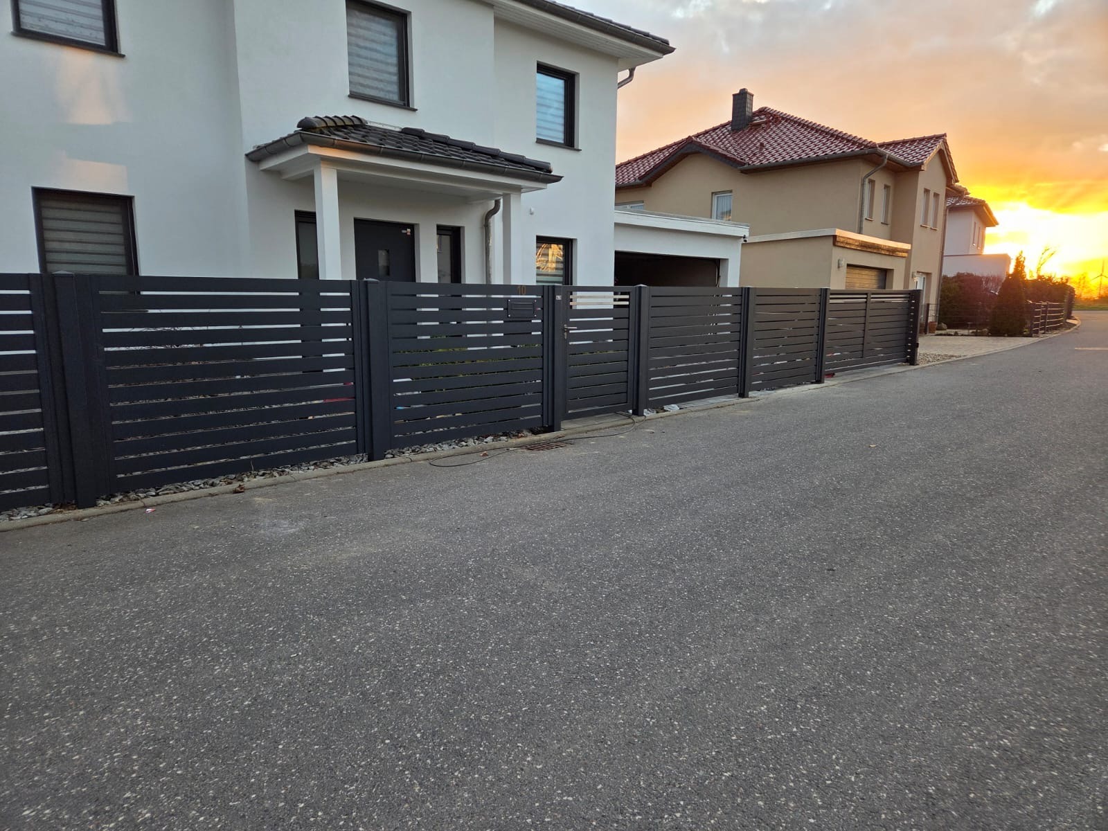 Nowoczesne, poziome ogrodzenie metalowe w kolorze antracytowym przed domem jednorodzinnym o minimalistycznej architekturze, widok z poziomu ulicy o zmierzchu.