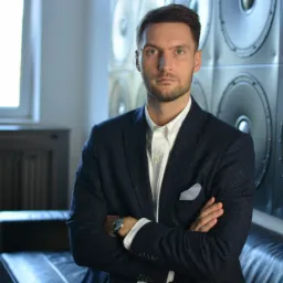 Mężczyzna w ciemnej marynarce, białej koszuli i zegarku na tle ściany z głośnikami i skórzanej sofy