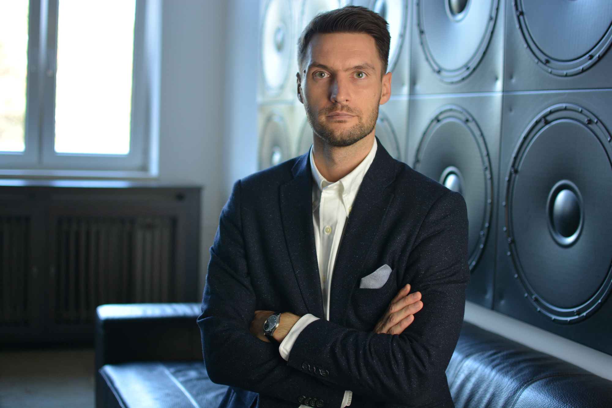 Mężczyzna w ciemnej marynarce, białej koszuli i zegarku na tle ściany z głośnikami i skórzanej sofy