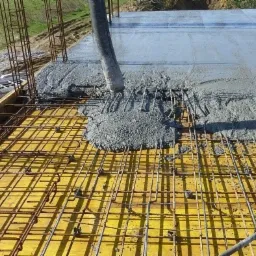 Wylewanie betonu na zbrojenie fundamentu z użyciem pompy, widoczny żółty szalunek i wystające pręty zbrojeniowe.