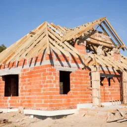 Surowy, ceglany dom w trakcie budowy z widoczną drewnianą konstrukcją dachu i nieotynkowanymi ścianami, w słoneczny dzień.