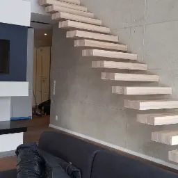 Nowoczesne schody drewniane wspornikowe zamontowane przy ścianie z betonu architektonicznego, obok ściana z kominkiem i telewizorem.