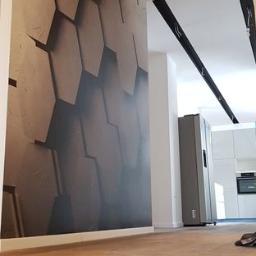 Przemysław Byszewski - Nowoczesne wnętrze z fototapetą o geometrycznym wzorze plastra miodu na ścianie, drewniana podłoga, widoczna biała kuchnia w tle, oświetlenie punktowe na suficie.