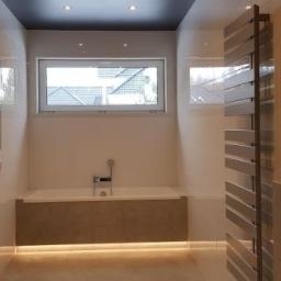 Przemysław Byszewski - Nowoczesna łazienka z białymi błyszczącymi płytkami, podświetloną wanną w zabudowie, grzejnikiem drabinkowym i oknami z widokiem na dachy sąsiednich domów.