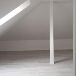 Przemysław Byszewski - Jasne, puste poddasze z drewnopodobną podłogą, skośnym oknem dachowym i białym słupem wspierającym strop.