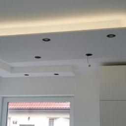 Przemysław Byszewski - Biały, podwieszany sufit z oświetleniem LED i czarnymi punktami świetlnymi w nowocześnie wykończonym wnętrzu, widoczne szafki kuchenne i okno.