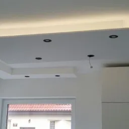 Biały, podwieszany sufit z oświetleniem LED i czarnymi punktami świetlnymi w nowocześnie wykończonym wnętrzu, widoczne szafki kuchenne i okno.