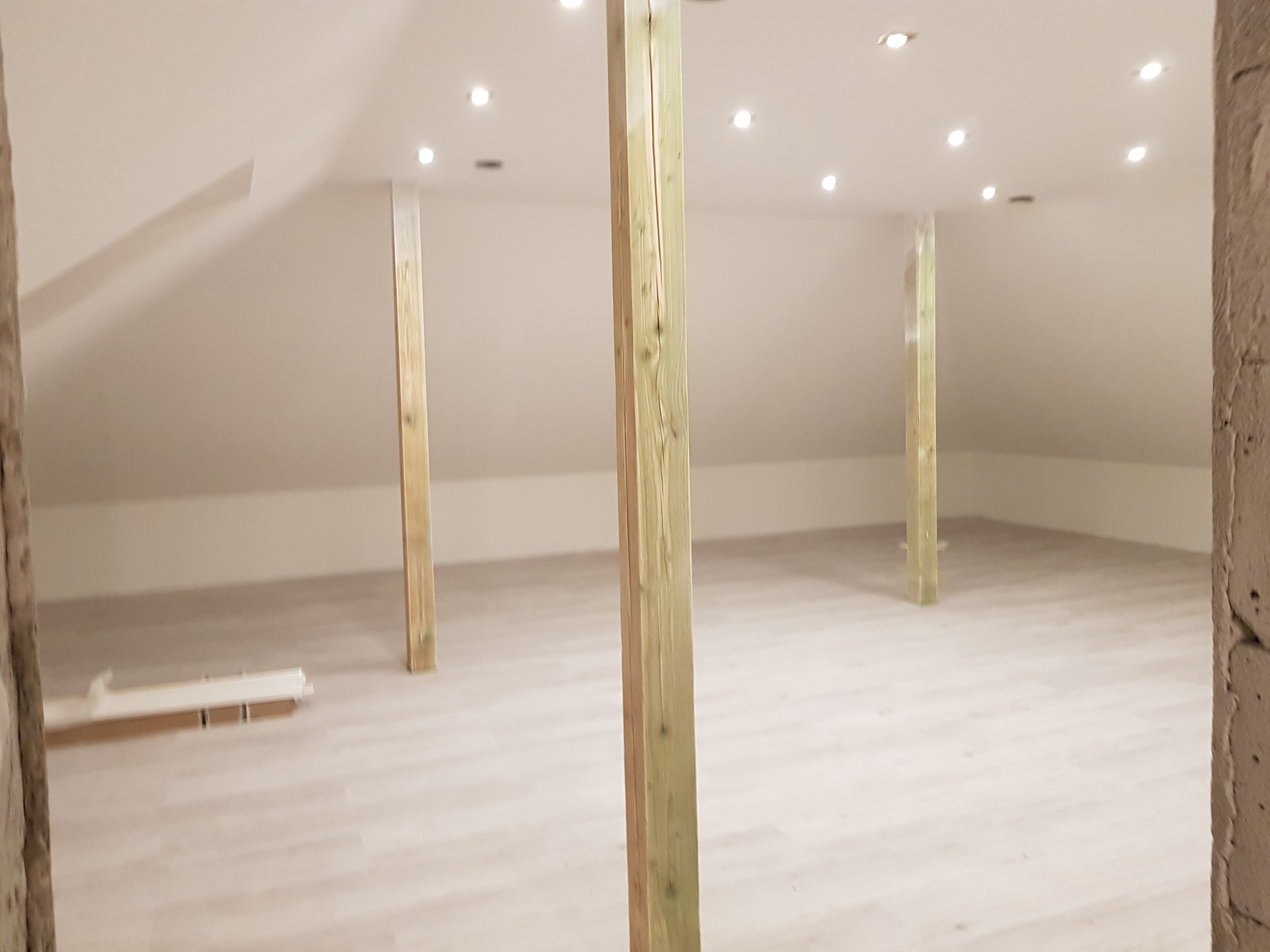 Poddasze z drewnianymi słupami konstrukcyjnymi i zamontowanym oświetleniem punktowym, widok na jasną podłogę i przygotowane listwy przypodłogowe.