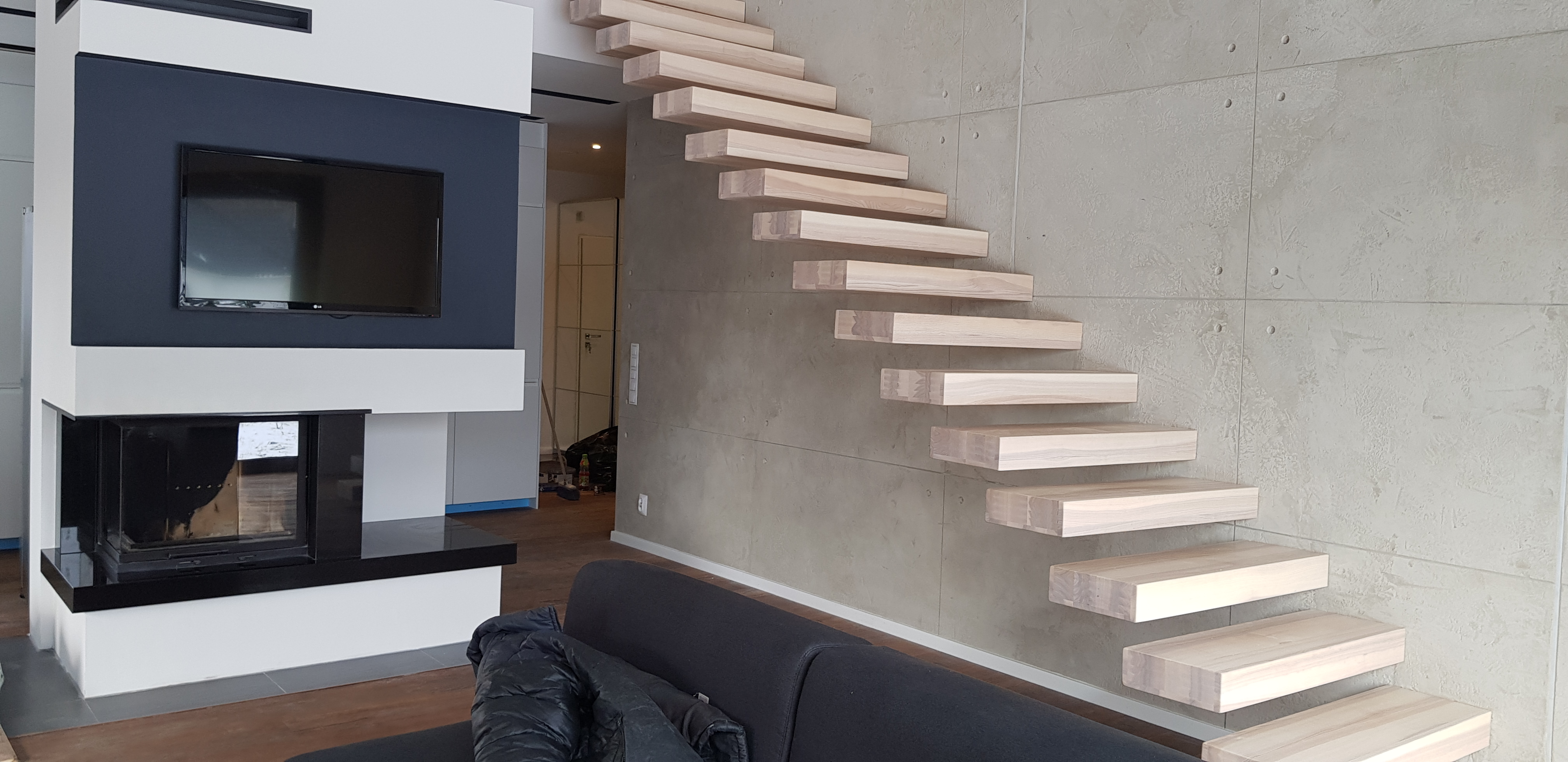 Nowoczesne schody drewniane wspornikowe zamontowane przy ścianie z betonu architektonicznego, obok ściana z kominkiem i telewizorem.