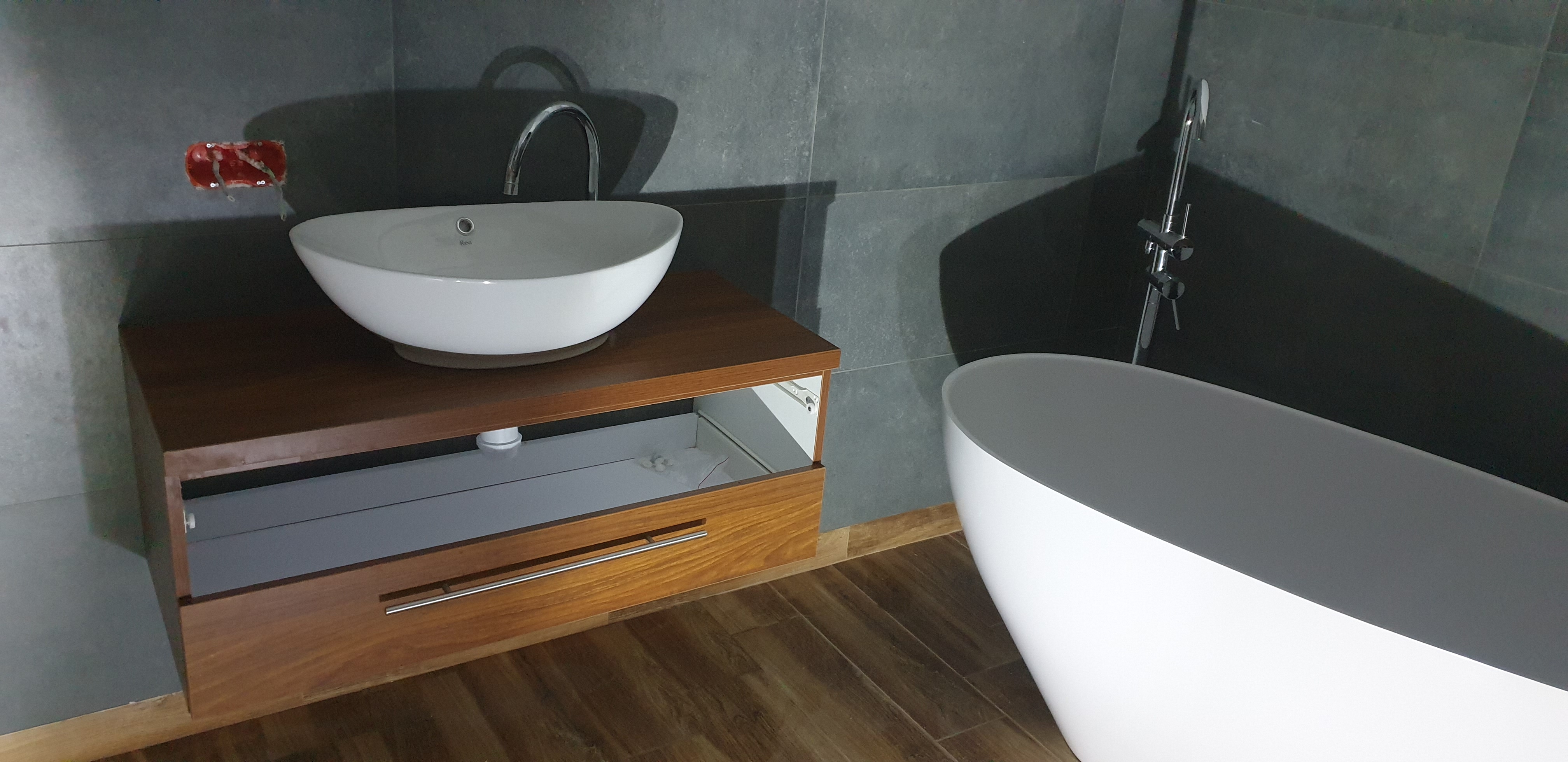 Umywalka nablatowa w kształcie misy na drewnianej szafce z wysuniętą szufladą, obok wolnostojąca wanna na tle szarych płytek. Widoczna instalacja elektryczna w ścianie.