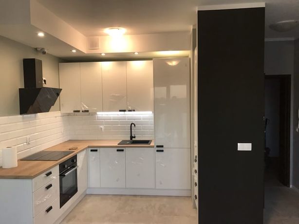 Nowoczesna kuchnia z białymi, lakierowanymi szafkami, drewnianym blatem i czarną armaturą, oświetlona punktowymi lampami sufitowymi, z widoczną czarną ścianą po prawej stronie.