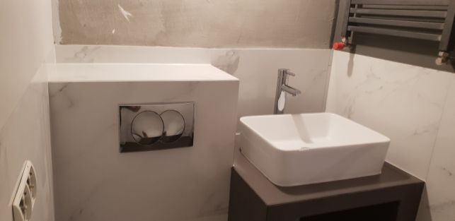 Wykończona łazienka z kwadratową białą umywalką, chromowaną baterią i podtynkową toaletą z przyciskiem spłukującym na tle białych płytek imitujących marmur, widoczne niedokończone szpachlowanie...