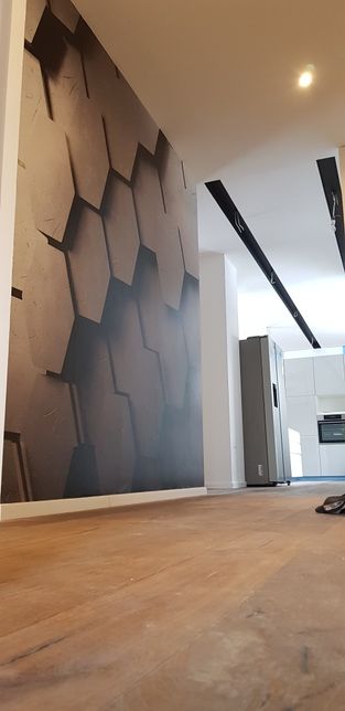 Nowoczesne wnętrze z fototapetą o geometrycznym wzorze plastra miodu na ścianie, drewniana podłoga, widoczna biała kuchnia w tle, oświetlenie punktowe na suficie.