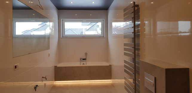 Nowoczesna łazienka z białymi błyszczącymi płytkami, podświetloną wanną w zabudowie, grzejnikiem drabinkowym i oknami z widokiem na dachy sąsiednich domów.
