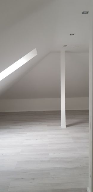 Jasne, puste poddasze z drewnopodobną podłogą, skośnym oknem dachowym i białym słupem wspierającym strop.