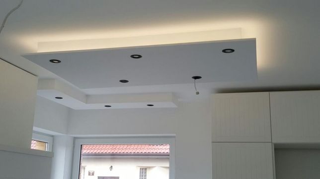 Biały, podwieszany sufit z oświetleniem LED i czarnymi punktami świetlnymi w nowocześnie wykończonym wnętrzu, widoczne szafki kuchenne i okno.