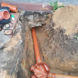 Pomarańczowe rury kanalizacyjne ułożone w wykopie ziemnym, widoczne połączenia kształtek, narzędzia i materiały na powierzchni, ściany wykopu z widocznymi korzeniami.