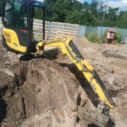Żółta minikoparka Yanmar SV22 w trakcie prac ziemnych, widoczny wykop i fragment ogrodzenia w tle.