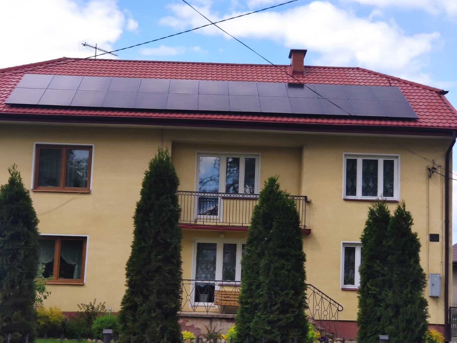 Dwupiętrowy dom z żółtą elewacją i czerwonym dachem, na którym zamontowane są panele fotowoltaiczne; na dachu widoczna antena telewizyjna.