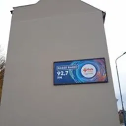 Ściana budynku z zamontowanym podświetlanym billboardem reklamowym radia 'Nasze Radio' 92.7 FM. Billboard z niebiesko-pomarańczową grafiką umieszczony na jasnej elewacji. Widoczny słup...