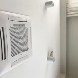 Kasetonowa jednostka wewnętrzna klimatyzacji zamontowana na białej ścianie, widoczne dwa cylindryczne reflektory i fragment lustra w srebrnej ramie.